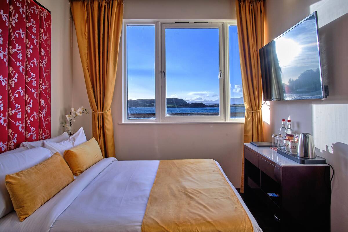 A hotel room with a bed, a desk with a TV, and a window with a view of the sea.