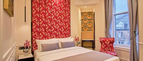 A hotel room with a large bed, a nightstand, a chair, a window with curtains, and a view of buildings outside.