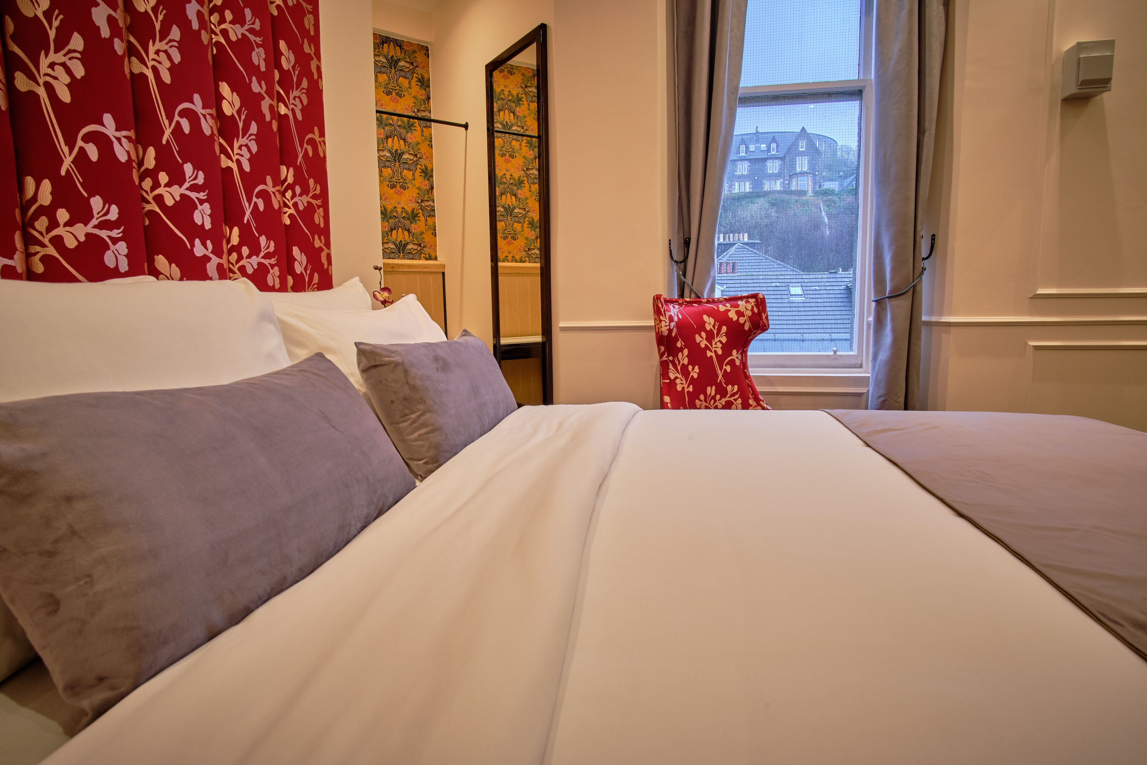 Ein Hotelzimmer mit einem Bett, einem roten Blumen-Kopfende, einem Spiegel und einem Fenster mit Blick auf Gebäude.