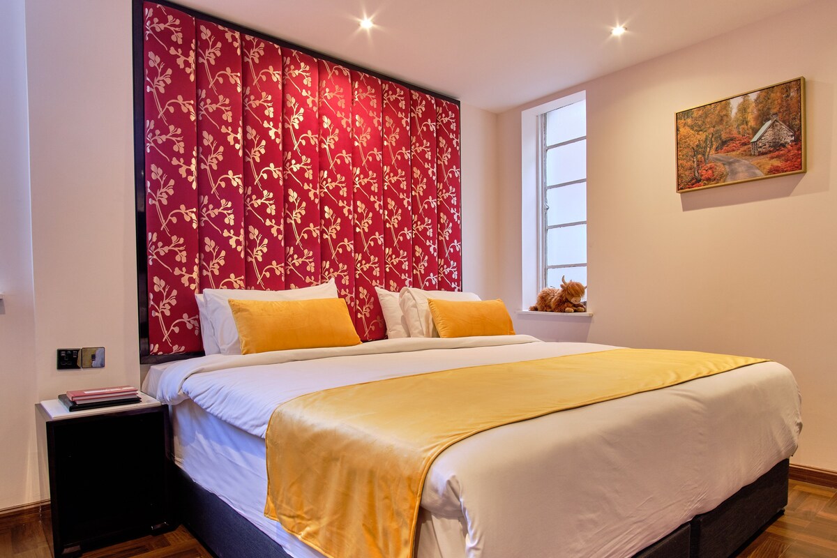 Una cama bien hecha con un plaid amarillo, un cabecero floral rojo y un cuadro en la pared.