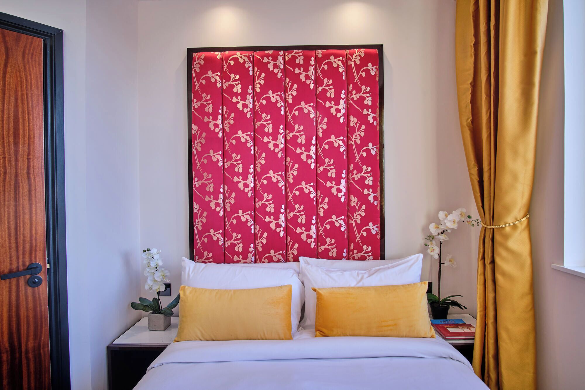 Un dormitorio con cama, dos almohadas, una mesita de noche con una planta y un cabecero floral rojo.