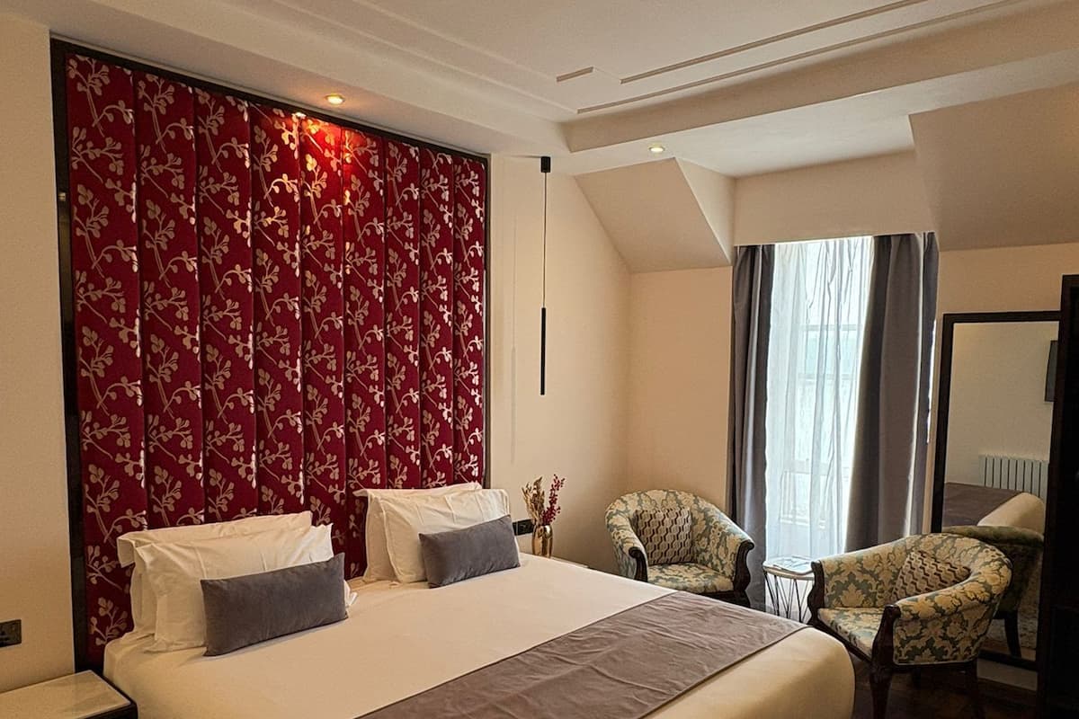 A spacious bedroom with a large bed, a decorative headboard, a seating area with two chairs, and a view of the city through a window.