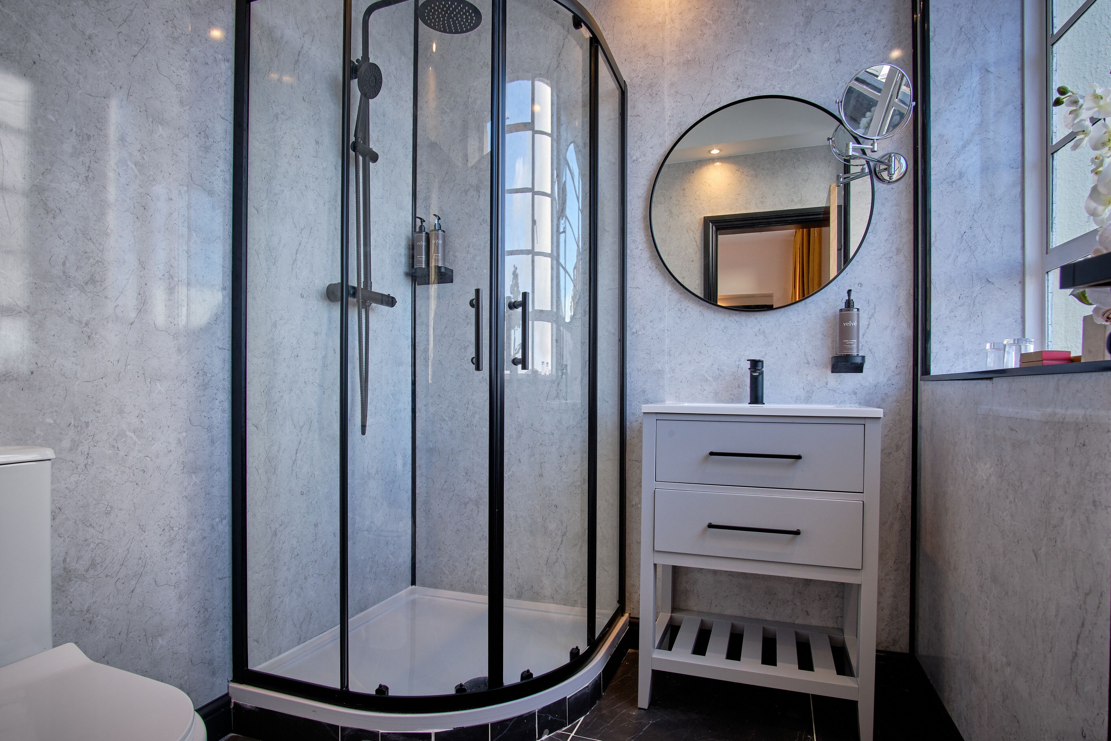 Une salle de bain moderne avec une douche à parois de verre, un miroir rond et un meuble-lavabo blanc doté de tiroirs.