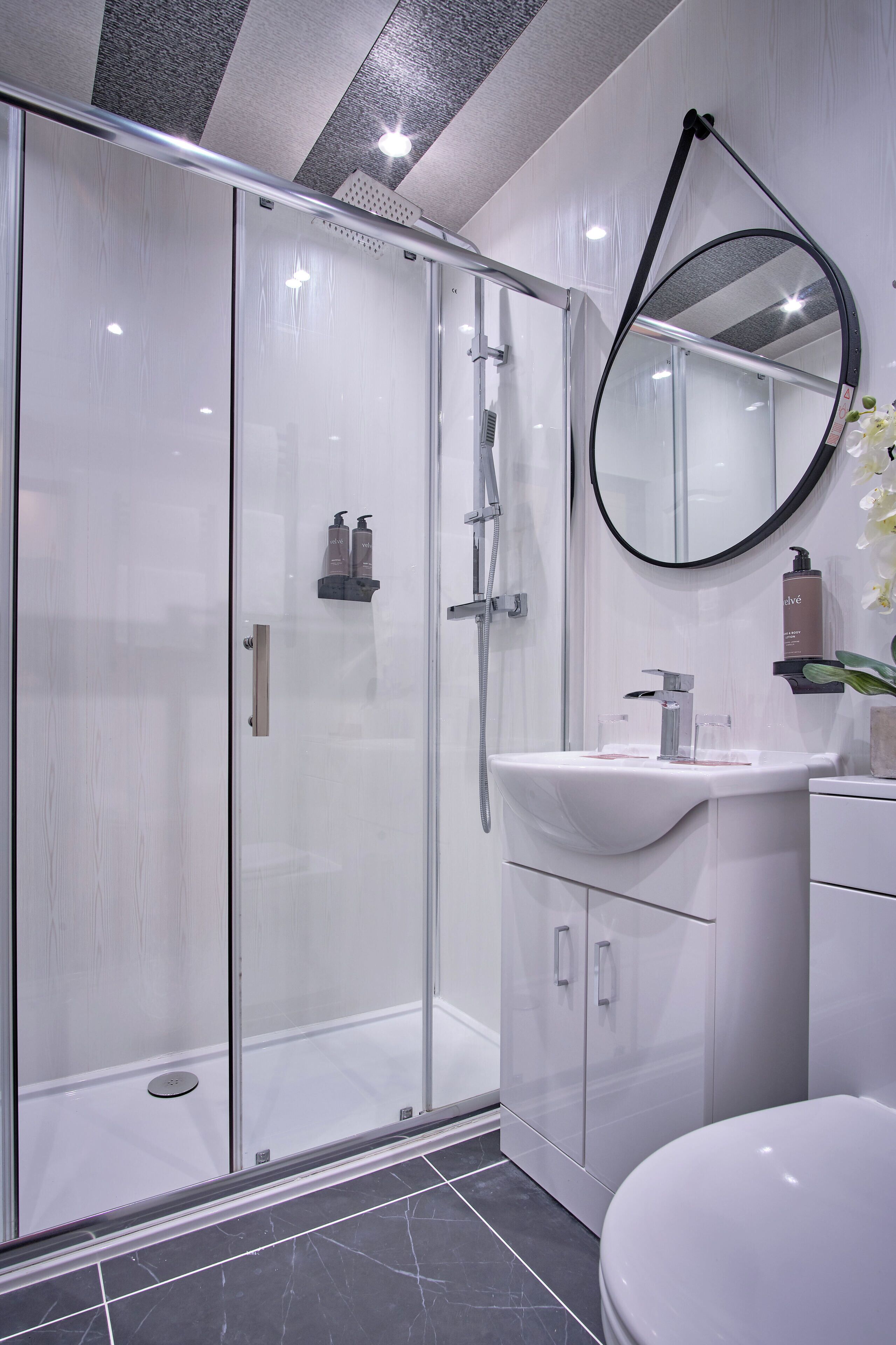 A modern bathroom with a glass-enclosed shower, a white sink, and a round mirror.