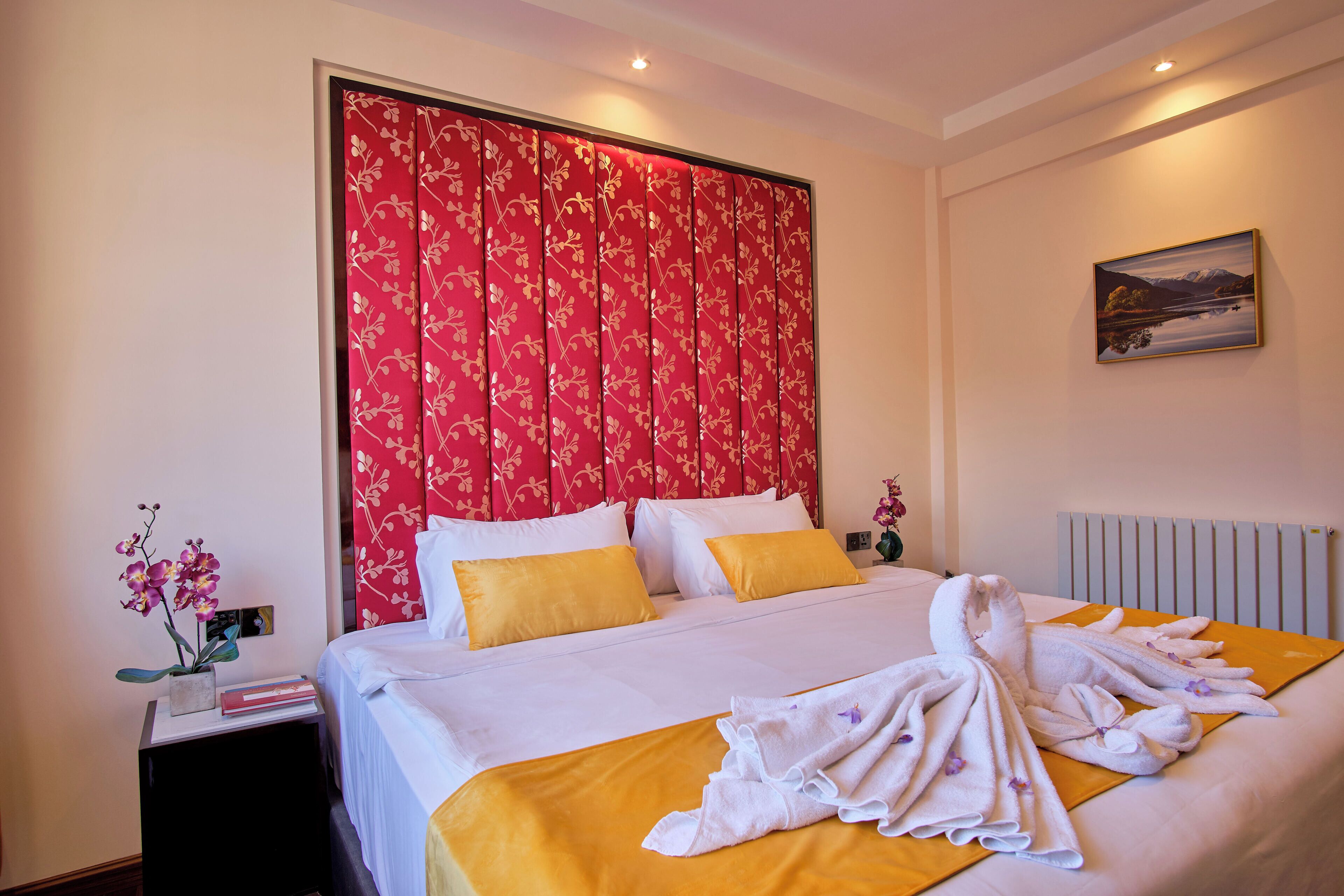 Een hotelkamer met een groot bed, een rood hoofdeinde met bloemenprint, wit beddengoed en een schilderij aan de muur.