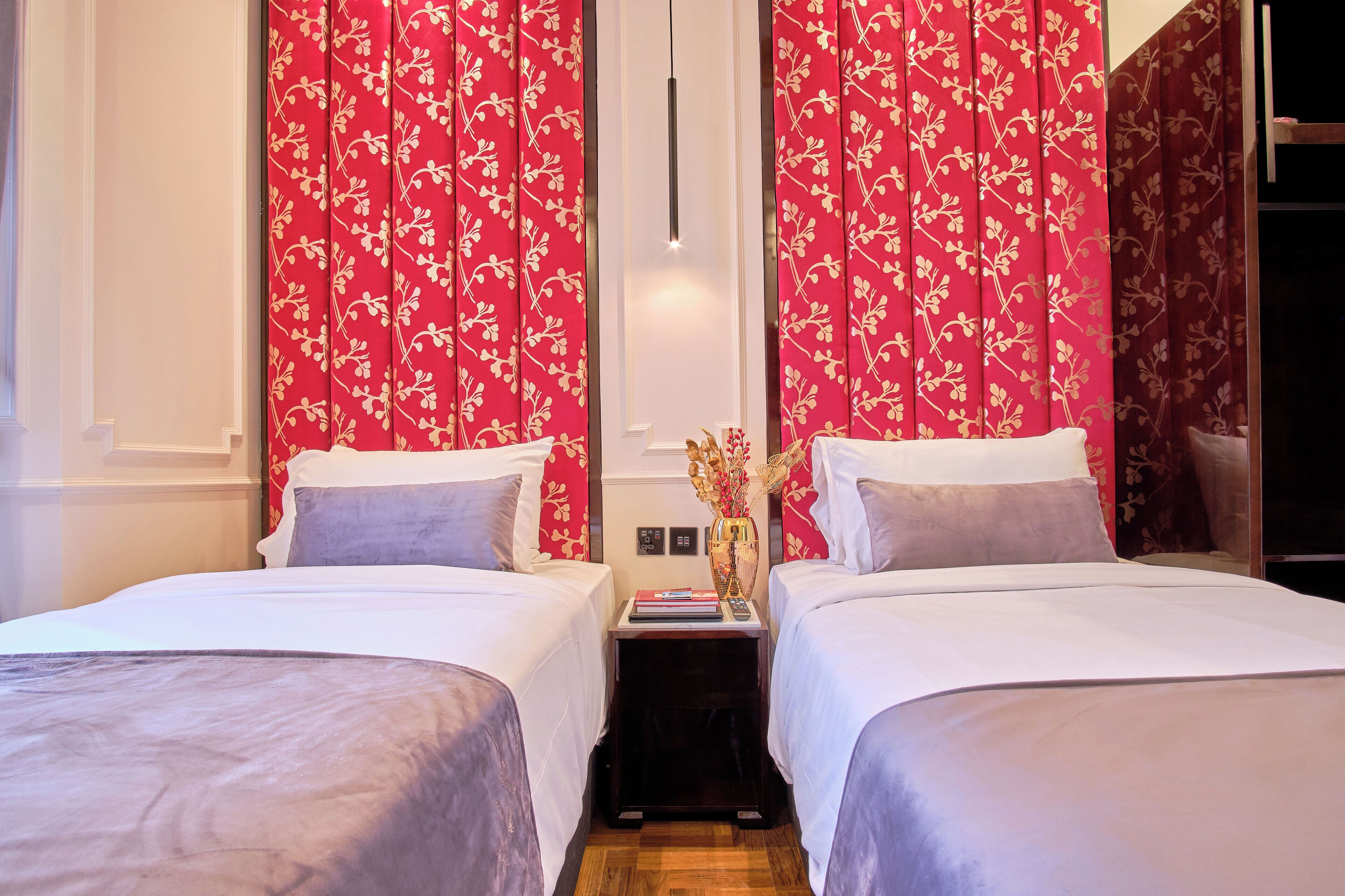 Habitación de hotel con dos camas, cortinas florales rojas, una mesita de noche con un florero y una lámpara moderna.