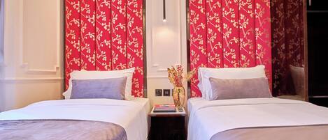 Habitación de hotel con dos camas, cortinas florales rojas, una mesita de noche con un florero y una lámpara moderna.