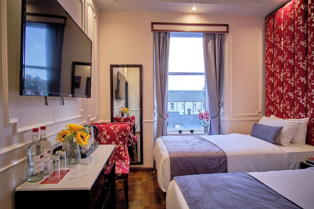 A hotel room with two beds, a TV, a desk with a vase of flowers, and a window with curtains.