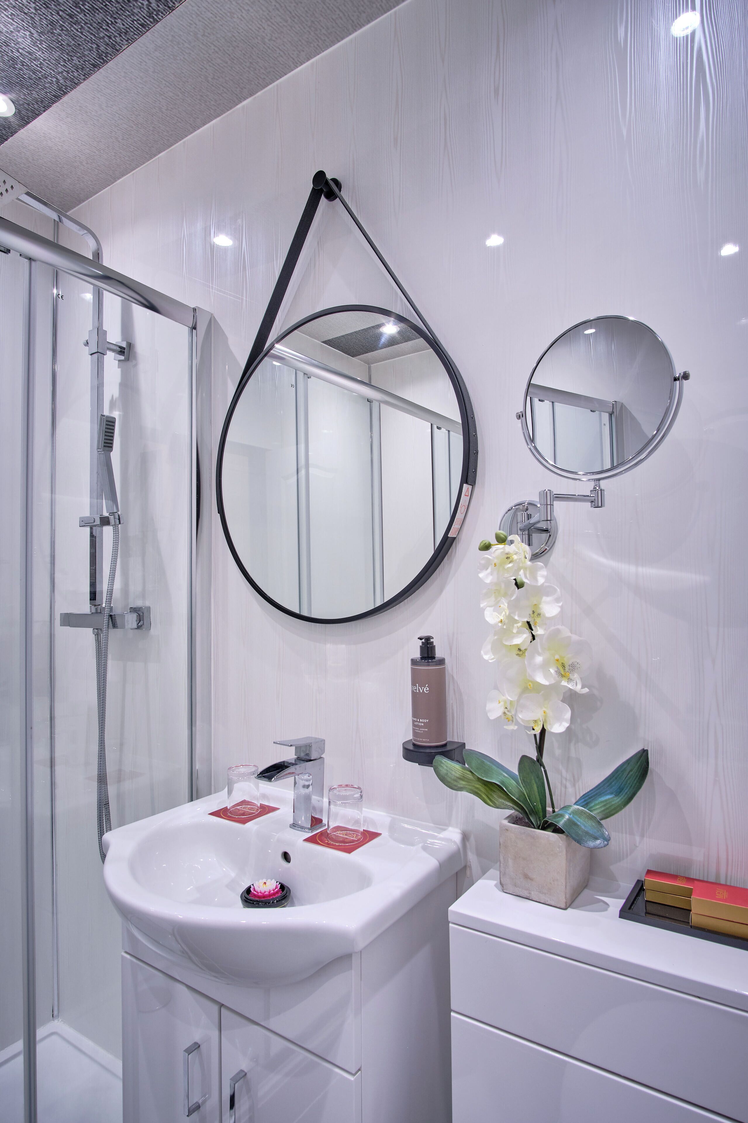 Une salle de bain moderne avec une douche à l’italienne en verre, un lavabo blanc doté d’un seul robinet et deux miroirs ronds fixés au mur.