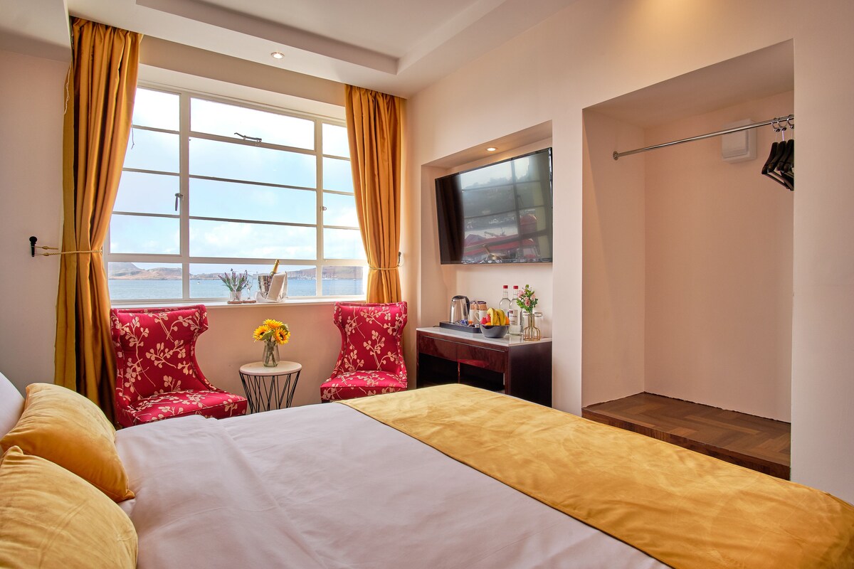 A hotel room with a bed, two red floral chairs, a small table with a vase of flowers, a desk with a TV, and a window with curtains.