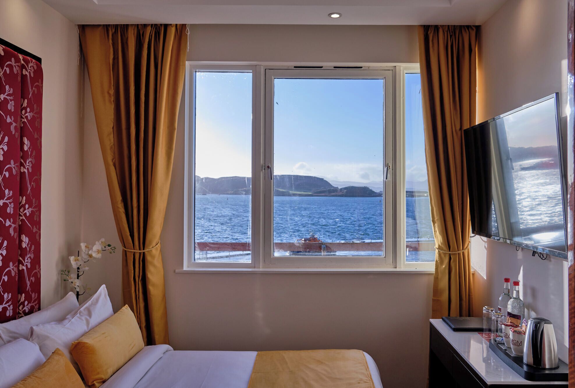 A hotel room with a bed, a desk with a laptop, a TV, and a window with a view of the sea and hills.