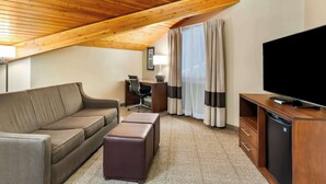 A hotel room with a wooden ceiling, a sofa, a desk with a chair, a television, and a window with curtains.