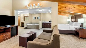 A hotel room with a large flat-screen TV, a sofa, a bed, a desk, and a bathroom visible through an open doorway.