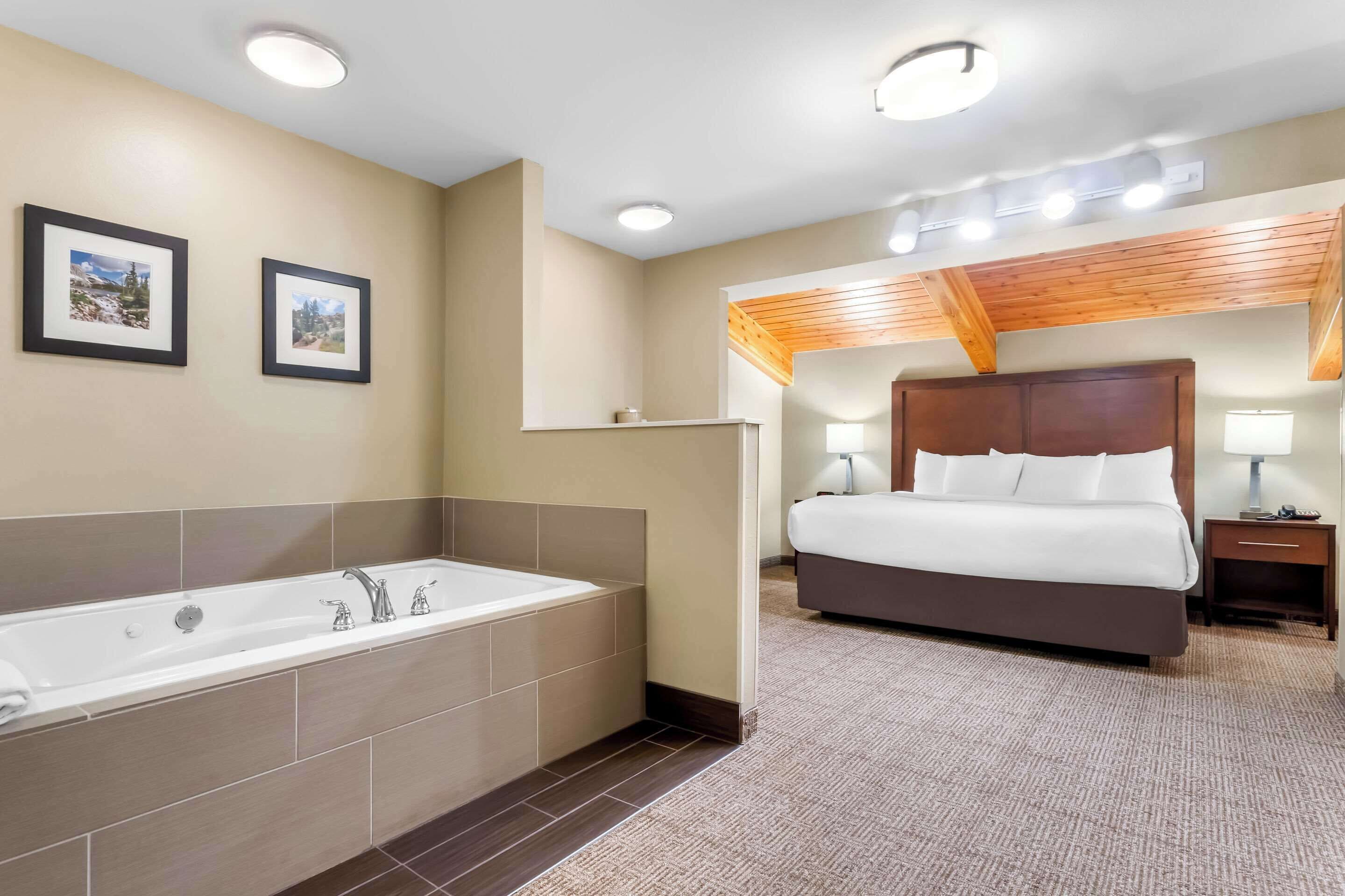 Une chambre d’hôtel avec une grande baignoire, un lit avec du linge de maison blanc, un plafond en bois et deux tableaux encadrés au mur.