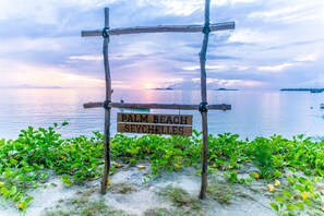 En träskylt med texten ”PALM BEACH SEYCHELLES” framför en strand med grönskande vegetation och ett lugnt hav.