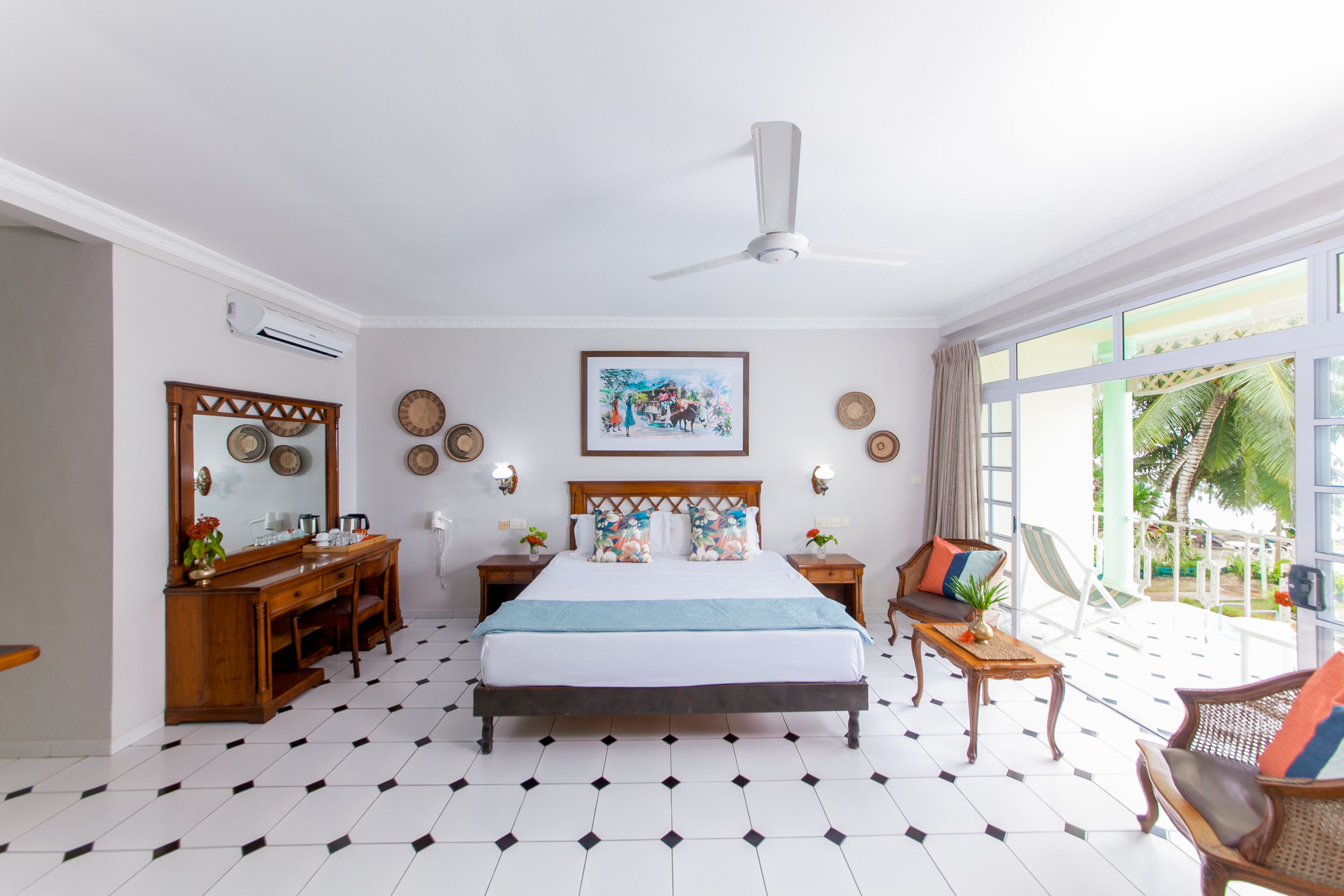A hotel room with a large bed, a wooden dresser, a desk, a chair, and a balcony with a view of palm trees.