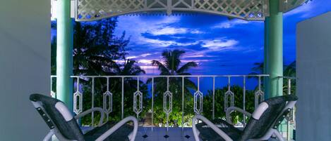 A balcony with two rocking chairs, a white railing, and a view of the sunset over palm trees.