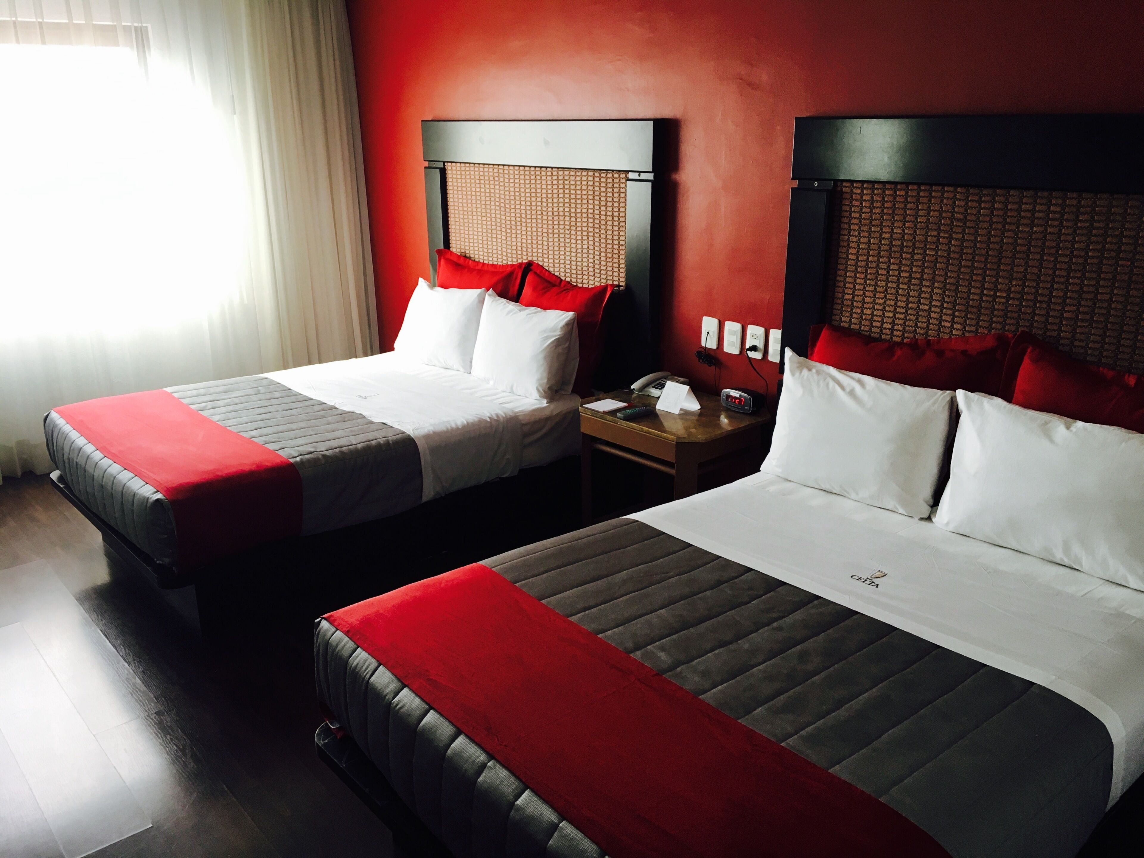 A hotel room with two beds, each with a red and white bedspread, a wooden nightstand with a telephone and a clock, and a red wall.