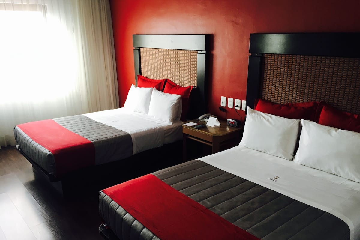 A hotel room with two beds, each with a red and white bedspread, a wooden nightstand with a telephone and a clock, and a red wall.