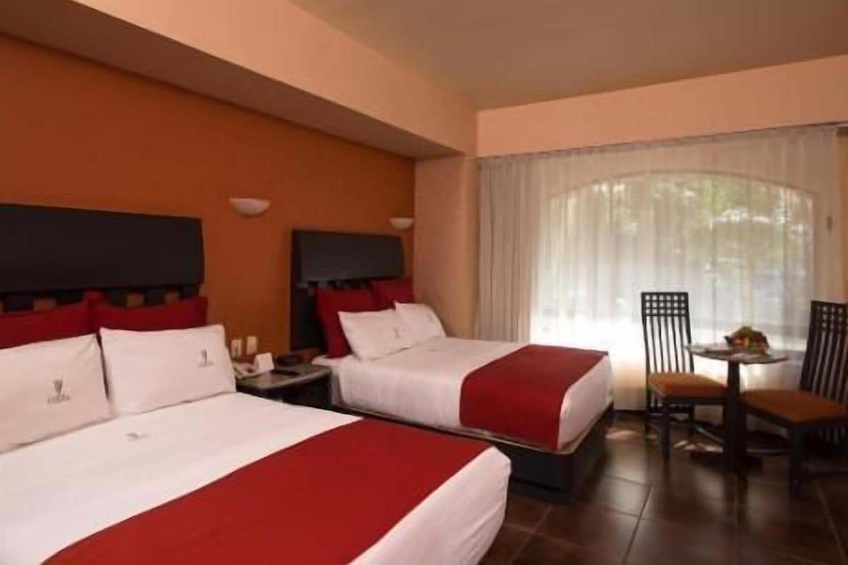 Habitación de hotel con dos camas, un televisor, una mesita con sillas y un ventanal grande con cortinas.