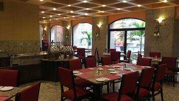 Un restaurante con sillas y mesas rojas, una zona de cocina y ventanas amplias que dan a vistas de palmeras.