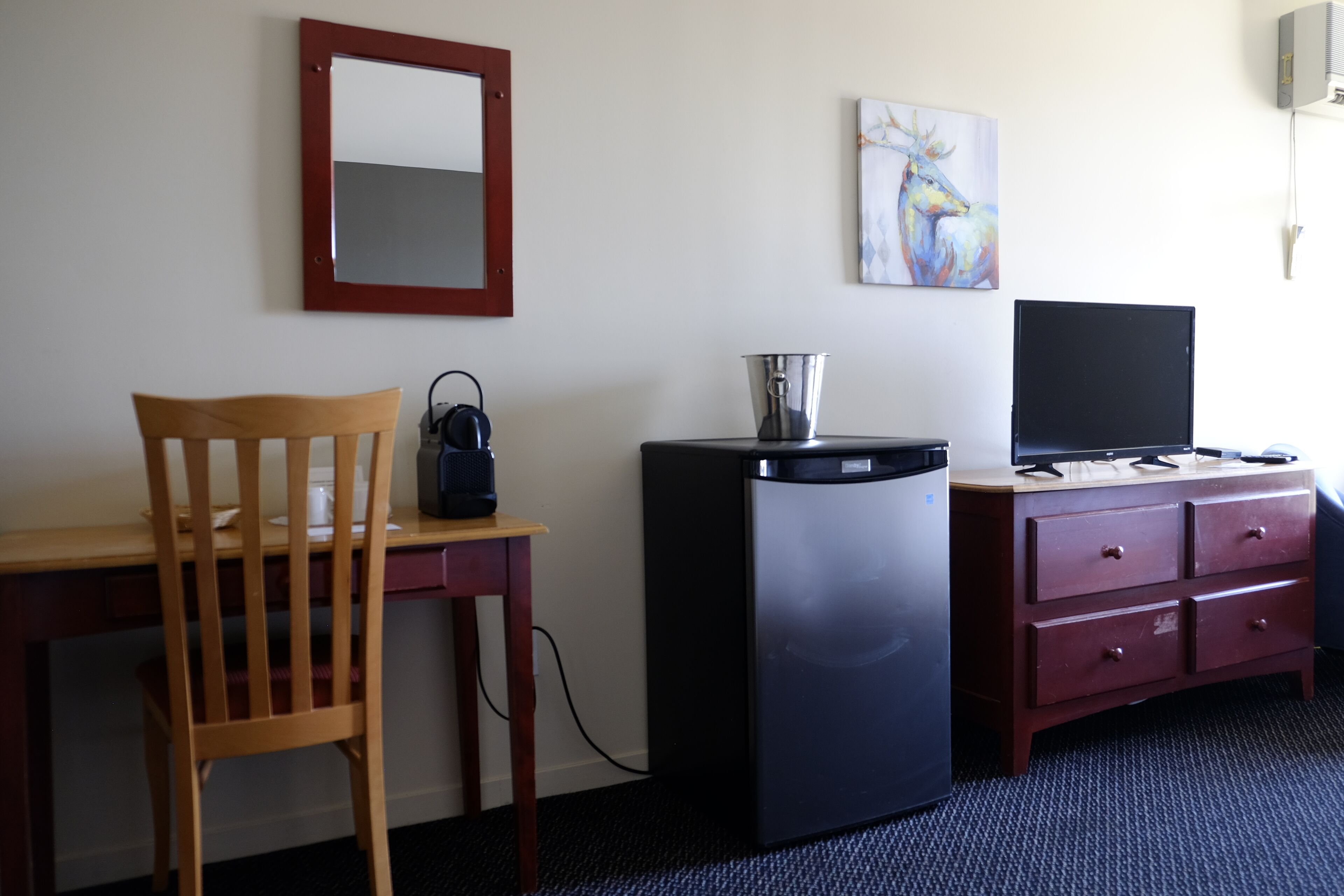 Een hotelkamer met een houten stoel, een bureau, een zwarte koelkast, een televisie op een houten dressoir, een schilderij aan de muur en een spiegel.