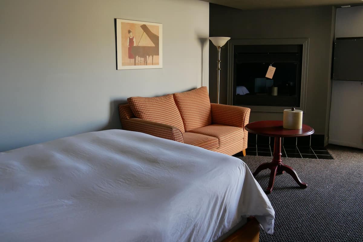 Una habitación de hotel con una cama, un sofá a cuadros, una mesita redonda pequeña, una lámpara y una ventana con vistas al exterior.