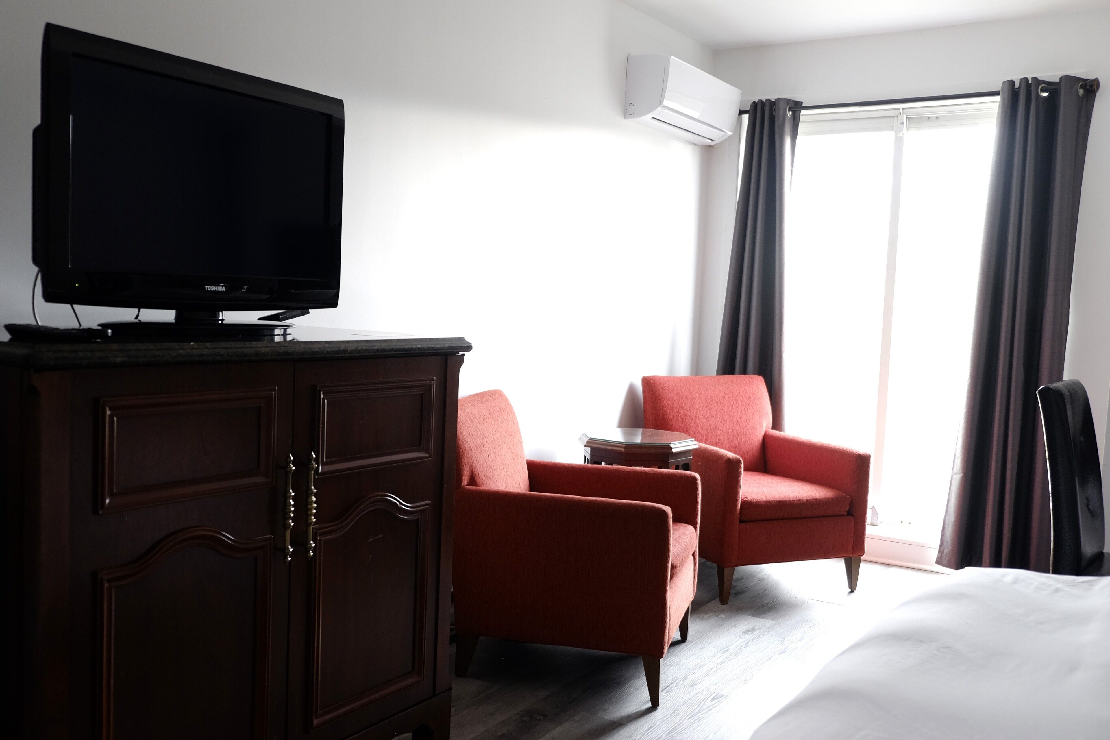A hotel room with a flat-screen TV on a cabinet, two red armchairs, a small round table, a bed, and a large window with curtains.