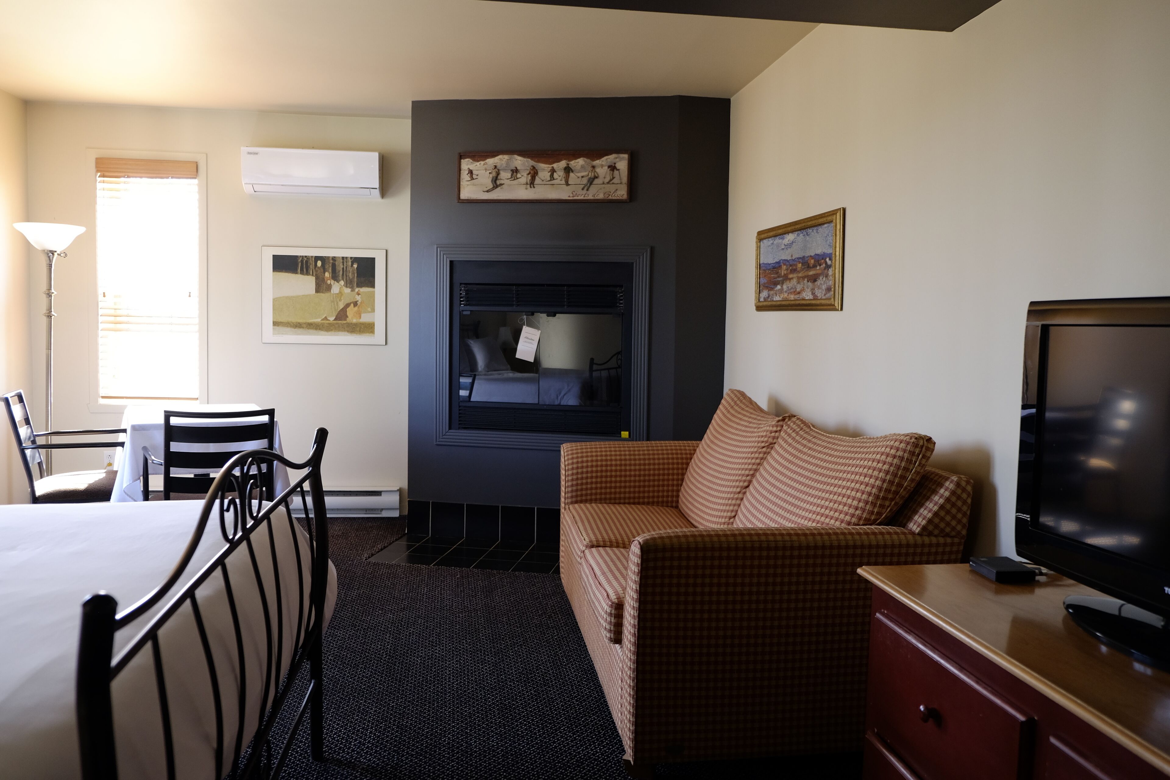 A hotel room with a bed, a checkered sofa, a television, a fireplace, and artwork on the walls.