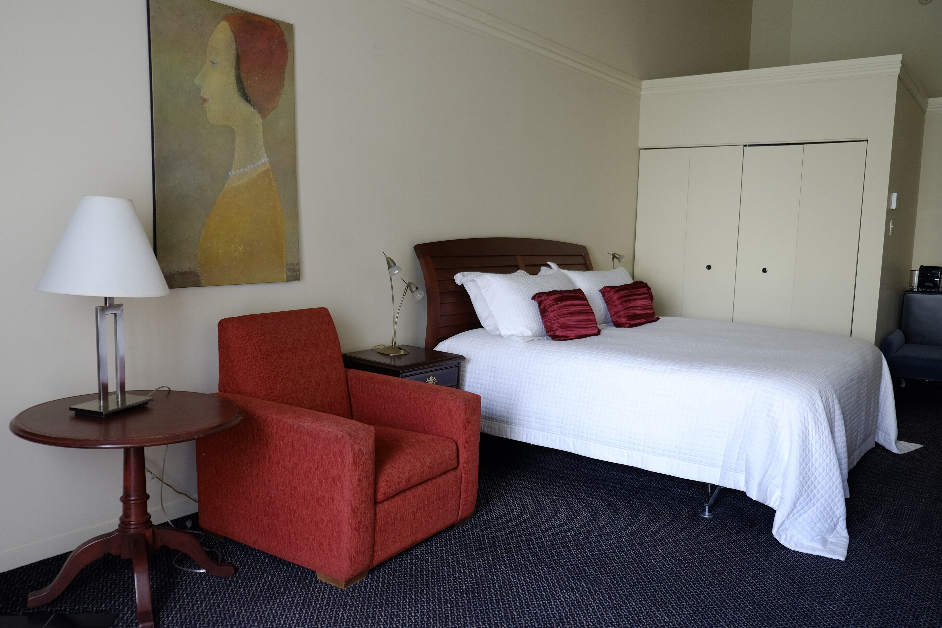A hotel room with a bed, a red armchair, a lamp, a painting on the wall, and a wardrobe.