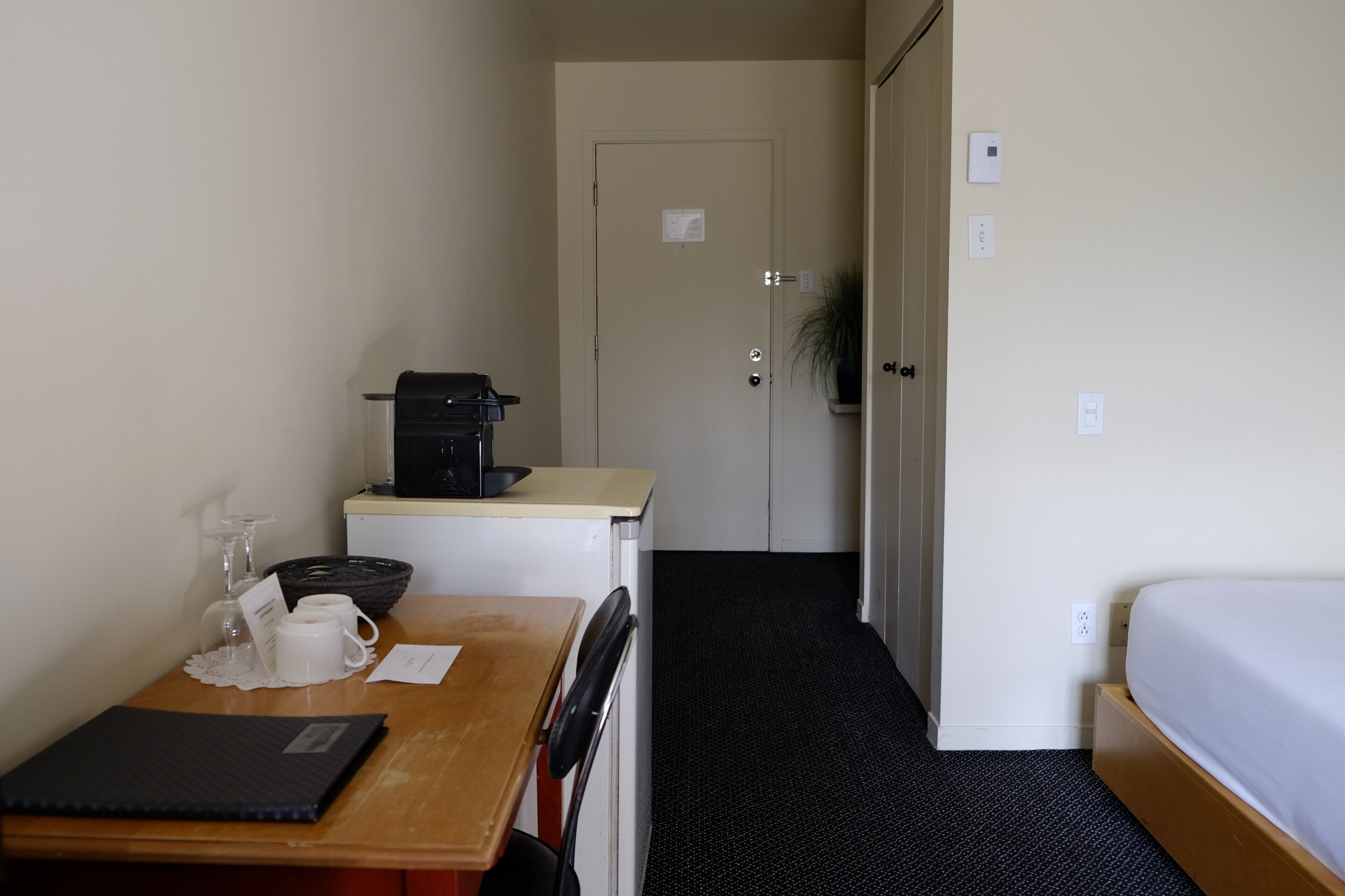 A small, compact room with a wooden desk, a black coffee maker, a white mug, and a glass on the desk. There is a bed with a white mattress and a black headboard. The room has a carpeted floor and a door leading to another room.