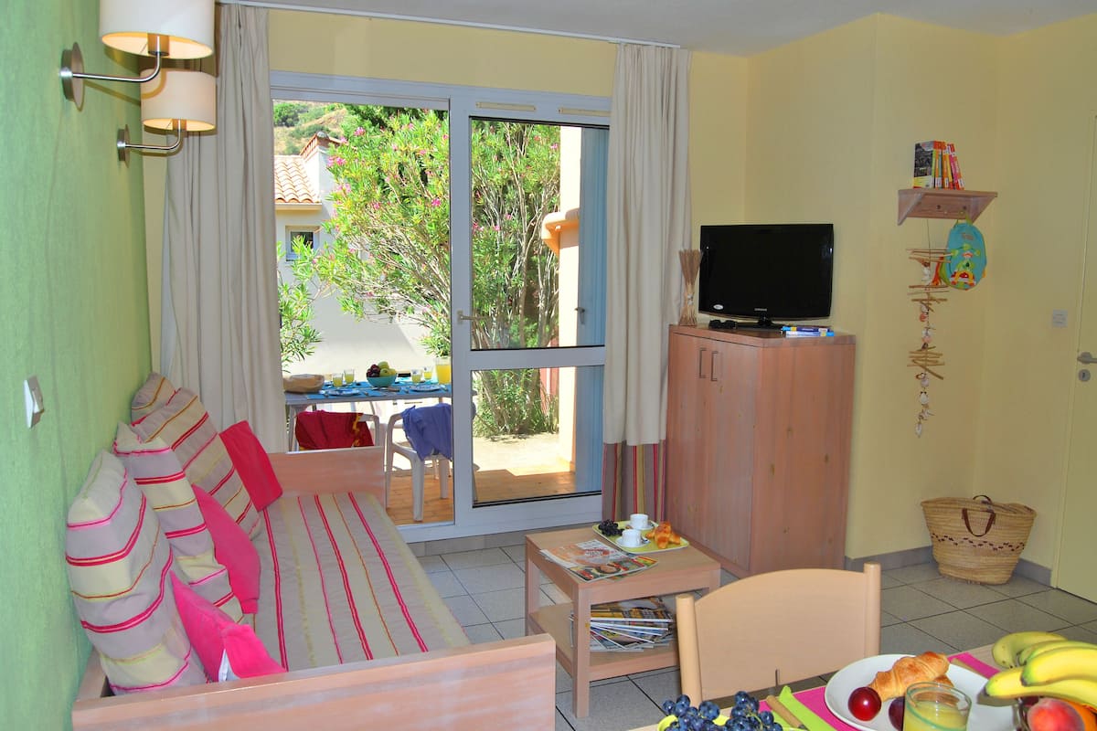 A room with a sofa, a television on a wooden cabinet, a dining table with a plate of fruit and a magazine, and a view of greenery outside through a sliding door.