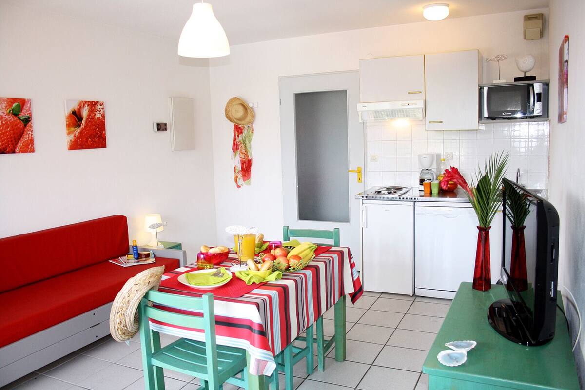 Un espace de vie compact comprenant un canapé rouge, une table à manger dressée, un coin cuisine avec un four à micro-ondes et un téléviseur posé sur un meuble.