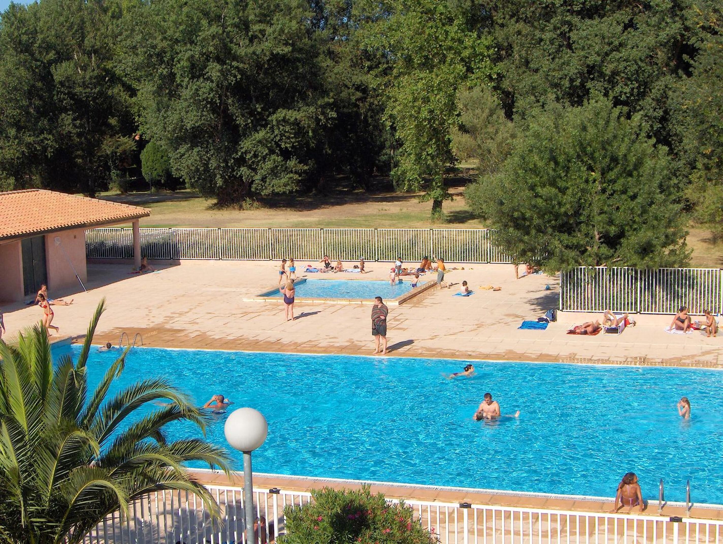 Ein Schwimmbad mit Schwimmern und Sonnenanliegern, umgeben von Bäumen und einem eingezäunten Bereich.