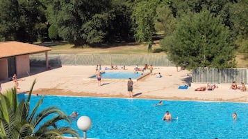 A swimming pool with people swimming and sunbathing, surrounded by trees and a fenced area.