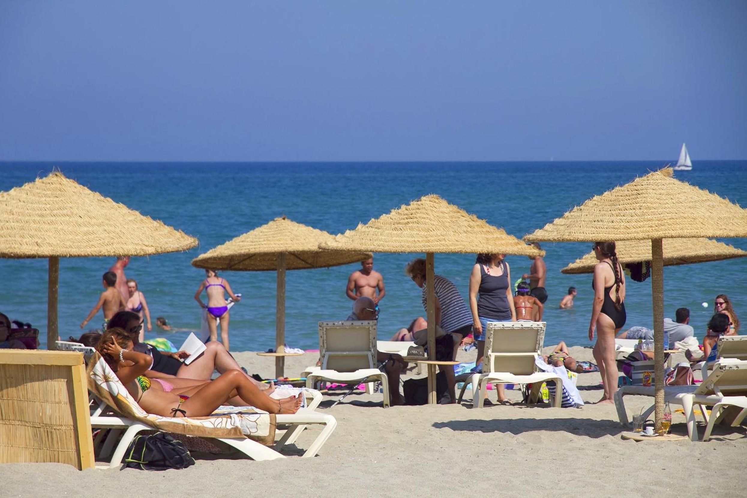 Ein Strand mit Liegestühlen und Reetsonnenschirmen, Personen, die sich sonnen und schwimmen, und ein Segelboot im Hintergrund.