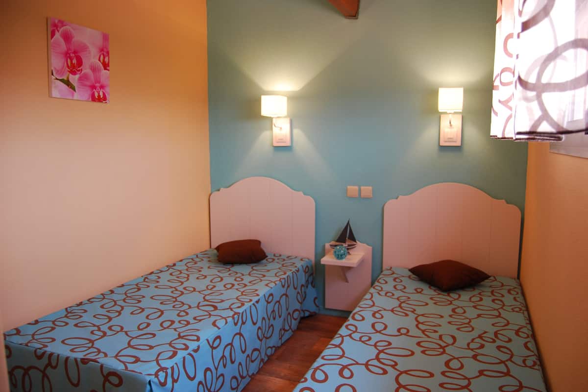 Ein kleines Zimmer mit zwei Betten, jeweils mit einem gemusterten Überwurf und einem Kissen. An den Wänden sind Leuchten angebracht und ein kleiner Nachttisch mit Lampe vorhanden.