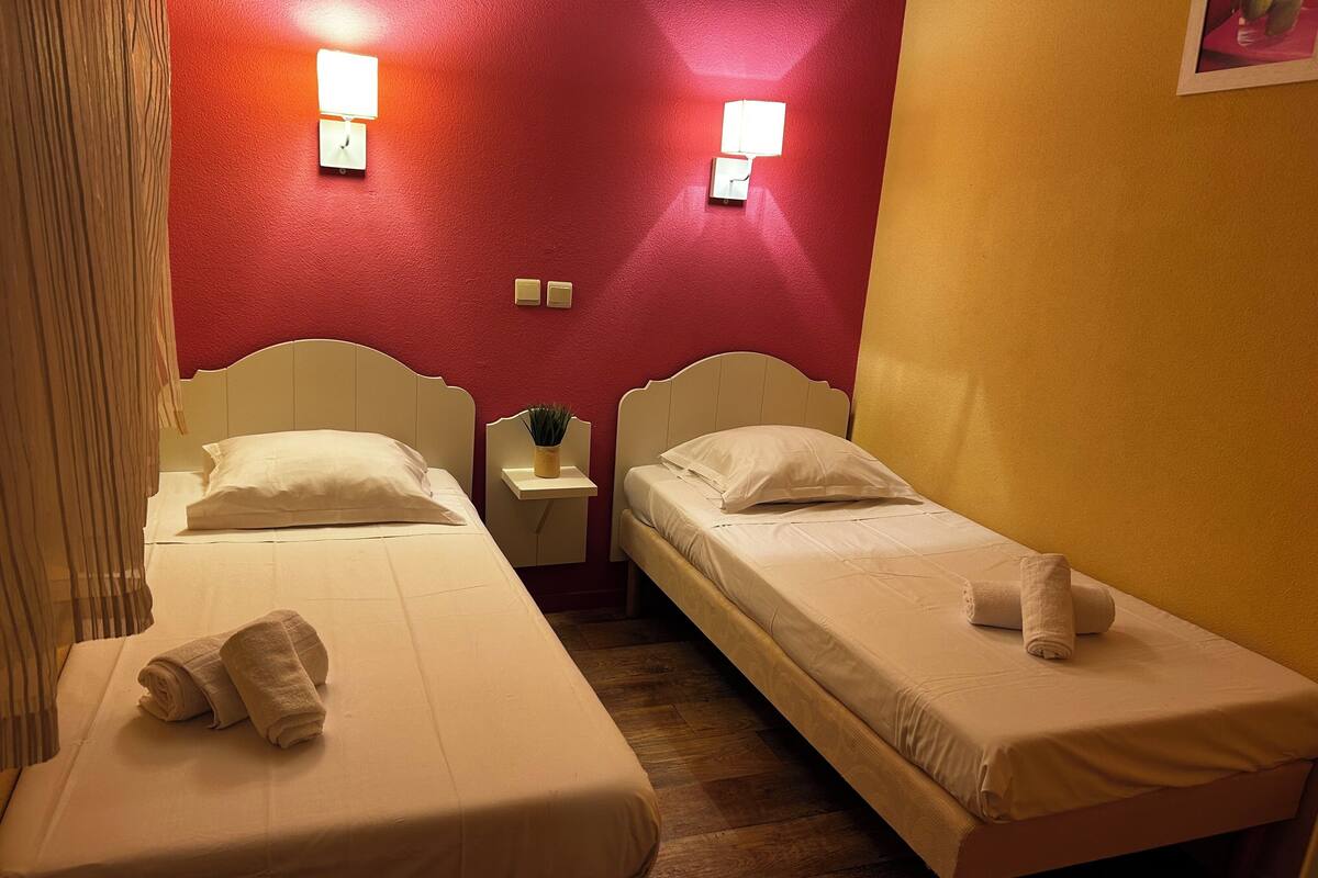 Ein Zimmer mit zwei Einzelbetten, jeweils mit einem Kopfkissen und einem zusammengerollten Handtuch. An den Nachttischen stehen Lampen und eine kleine Pflanze. Die Wände sind rot und gelb gestrichen.