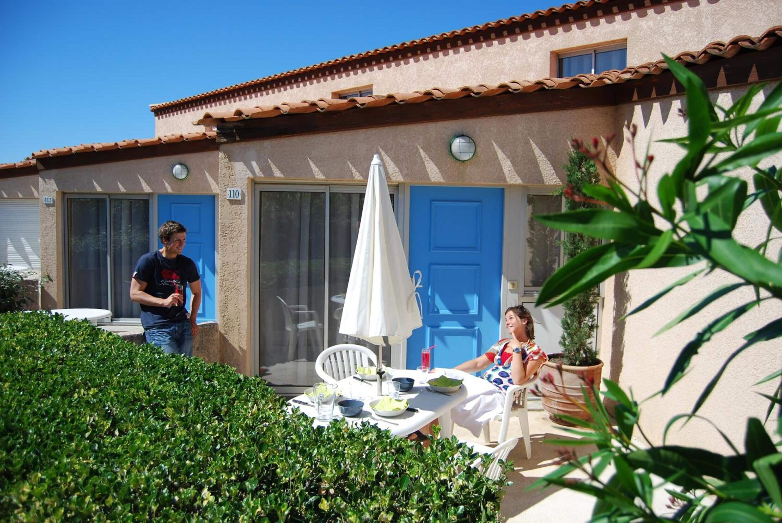Un patio con tavolo, sedie e un ombrellone bianco. Si vedono due porte, una blu e una bianca, e un'apertura finestrata.