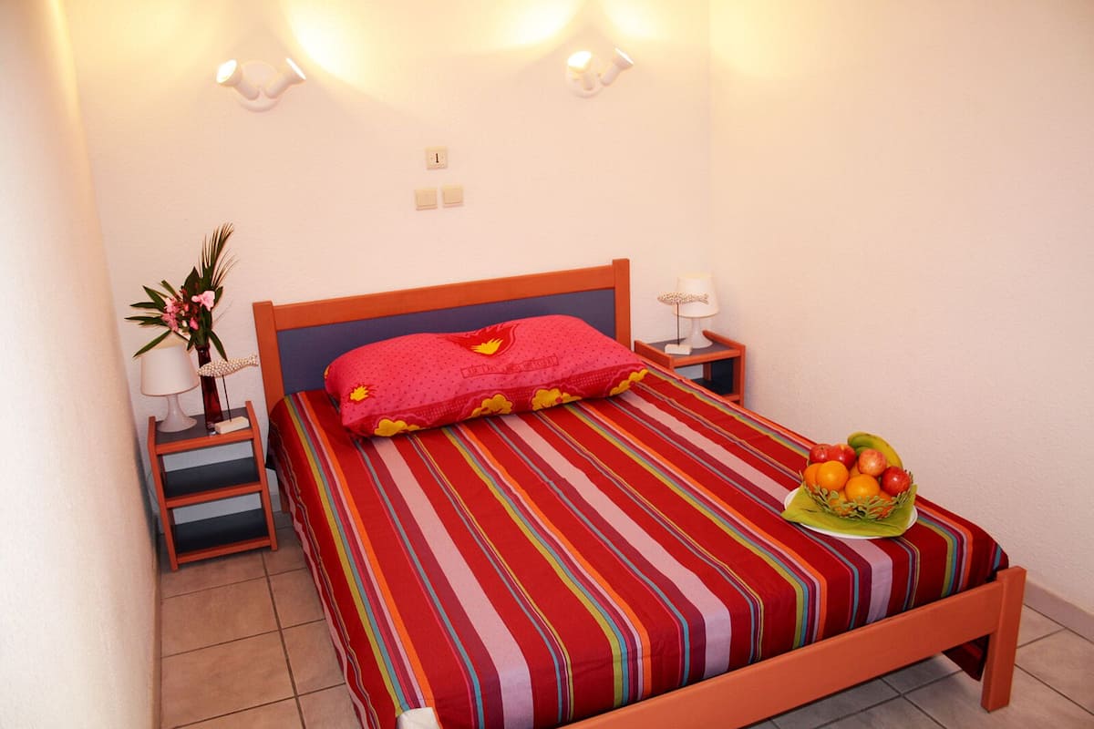 Ein Bett mit gestreiftem Bettbezug, einem roten Kissen und einem Obstkorb daneben. Zwei Nachttische mit Lampen und einer kleinen Pflanze sind ebenfalls vorhanden.