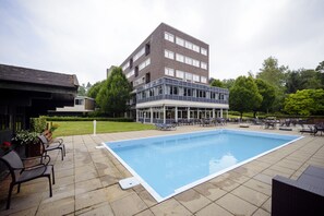 A rectangular outdoor swimming pool with a surrounding patio area, including chairs and tables, and a multi-story building in the background. - Fletcher Hotel-Restaurant Beekbergen - Apeldoorn (Beekbergen)