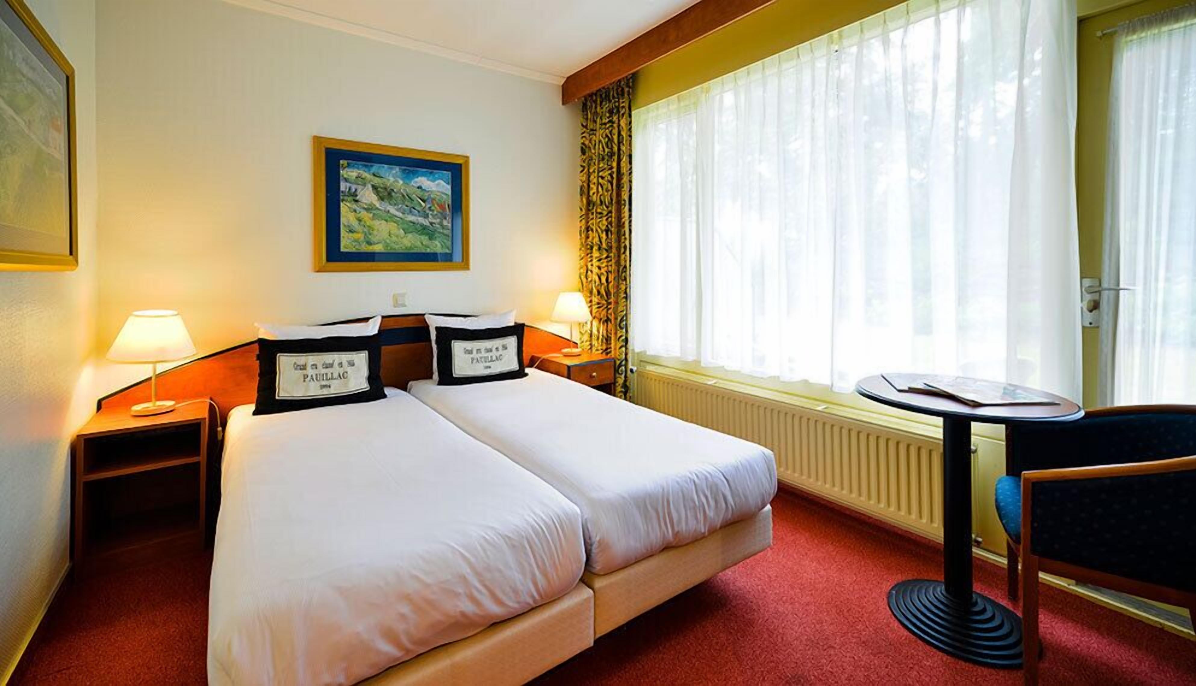 A hotel room with two beds, a painting on the wall, a window with curtains, a table, and a chair.