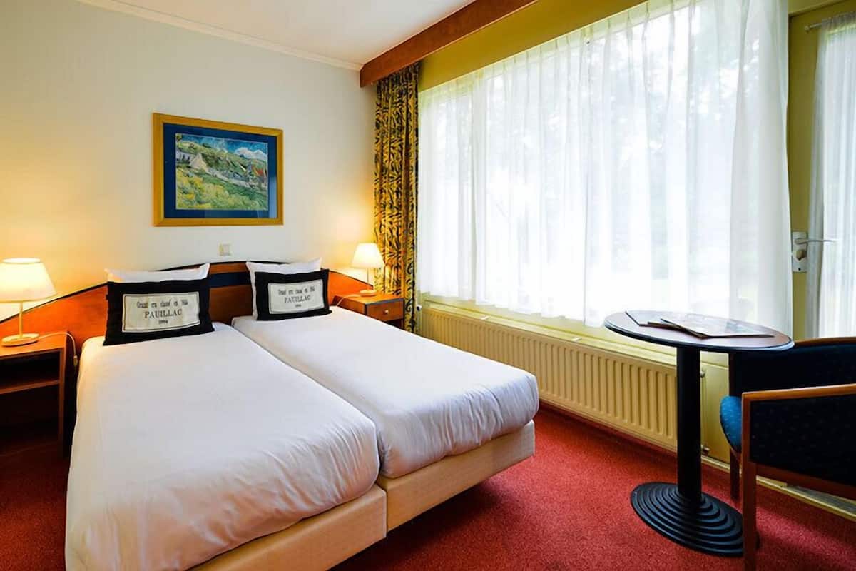 Ein Hotelzimmer mit zwei Betten, einem Bild an der Wand, einem Fenster mit Vorhängen, einem Tisch und einem Stuhl.