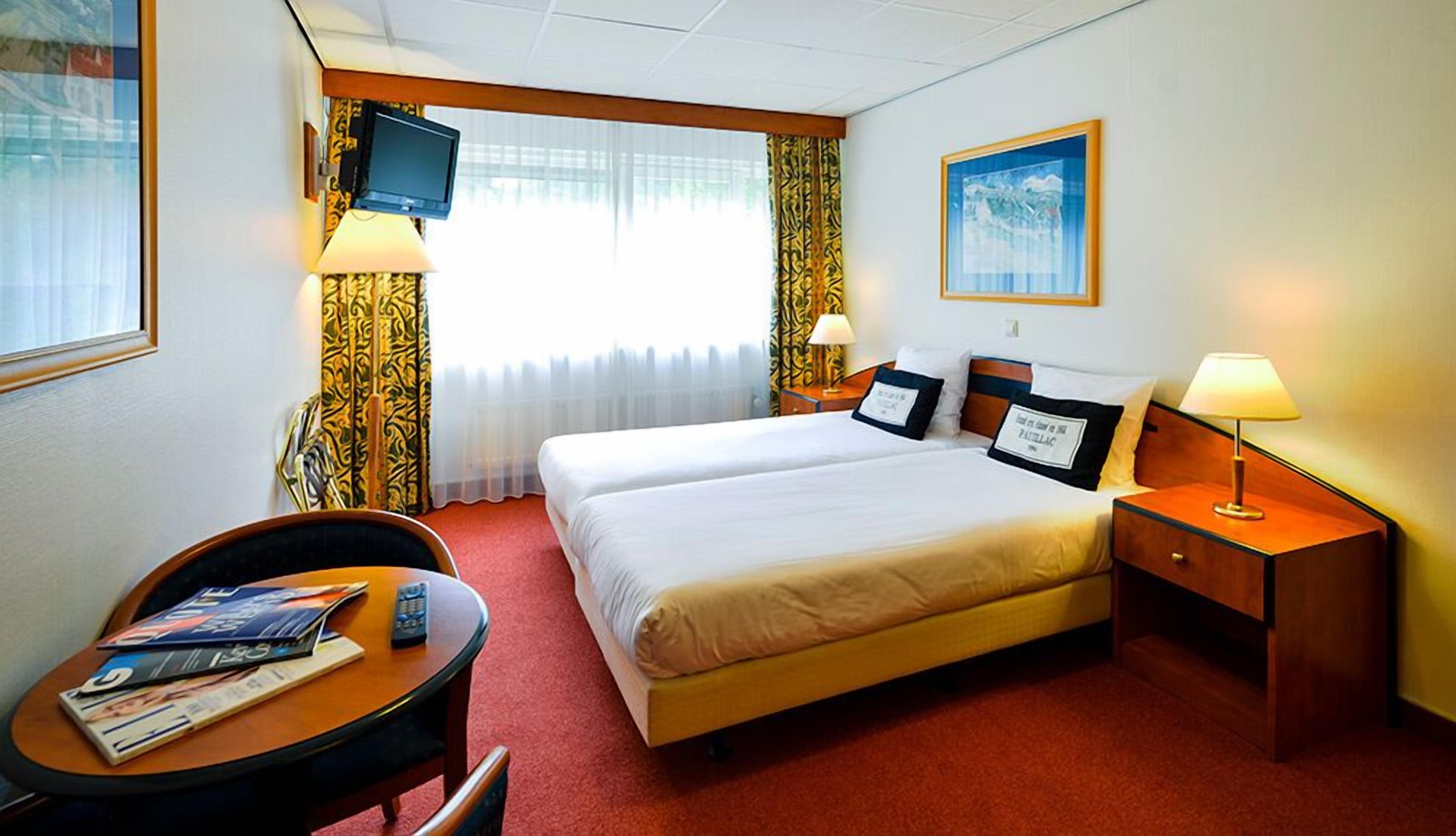 A hotel room with a double bed, two bedside tables, a TV mounted on the wall, a desk with a chair, and a window with curtains.