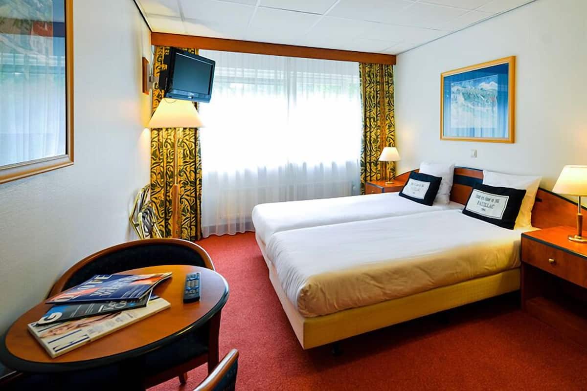 Ein Hotelzimmer mit zwei Betten, einem an der Wand montierten Fernseher, einem Schreibtisch mit Stuhl und einem Fenster mit Vorhängen.