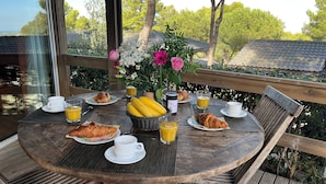 Ein Holztisch ist reichhaltig mit Frühstücksleckereien gedeckt, darunter Croissants, Saft und eine Blumenvase.