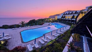 A pool area with lounge chairs and umbrellas, overlooking a body of water at sunset. - Caloura Hotel Resort (Lagoa)