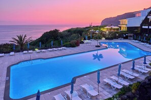 A pool with lounge chairs and umbrellas, surrounded by greenery and overlooking the ocean at sunset.
