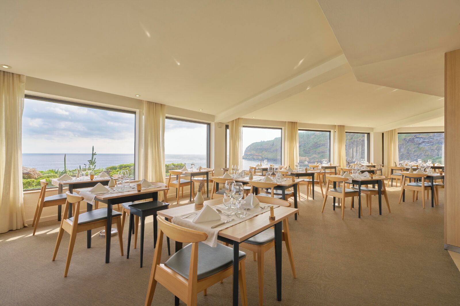 A restaurant with multiple tables and chairs, large windows offering a view of the sea and cliffs.
