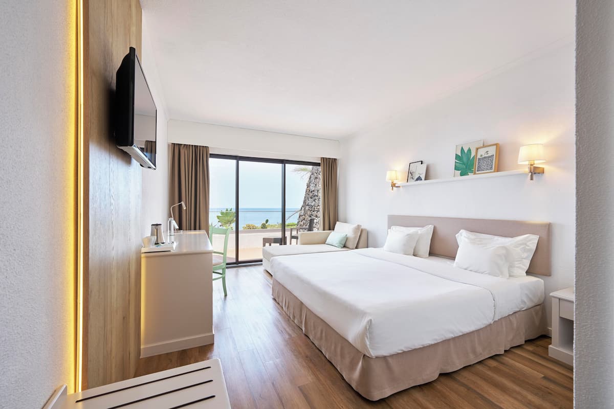A hotel room with a bed, bedside tables, a TV, a desk with a chair, a view of the sea, and a balcony.